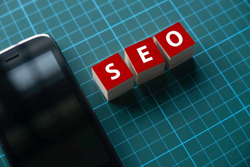 SEO-blokken met rode letters naast een smartphone op een digitaal raster, symbool voor zoekmachineoptimalisatie en mobiele vindbaarheid.